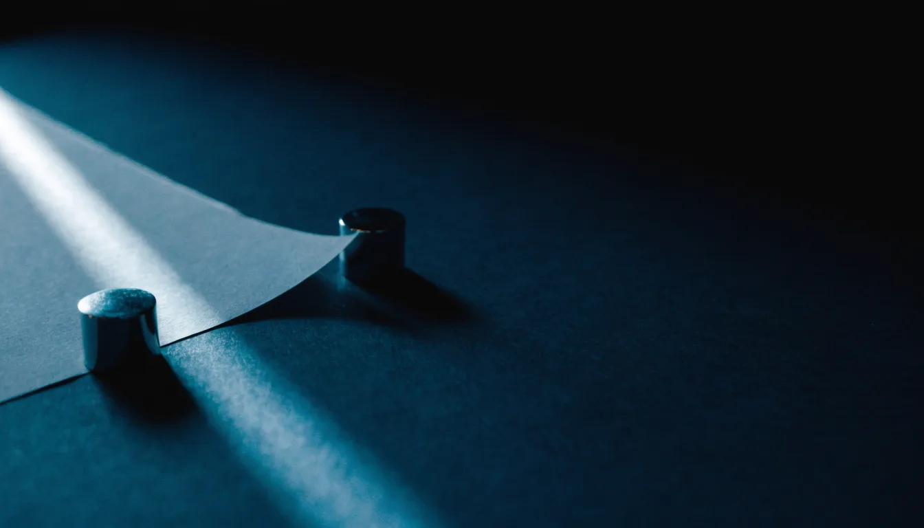A sheet of white paper pinned flat by two steel weights under cool cyan light, one corner lifting