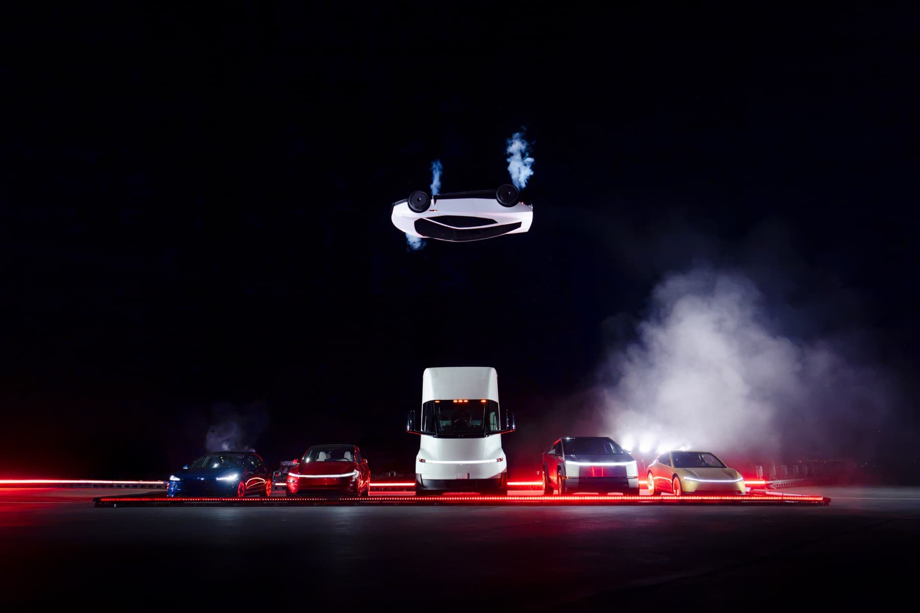 Inverted Roadster above a line of Tesla cars and a Semi at night.