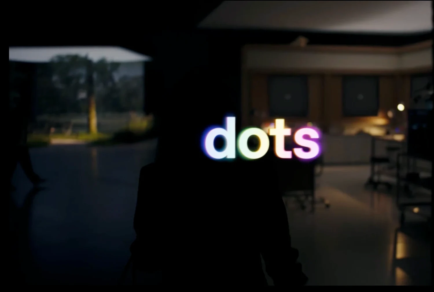 A dark, out-of-focus office interior seen through a doorway, with the word dots glowing in rainbow-coloured letters in the centre of the frame.