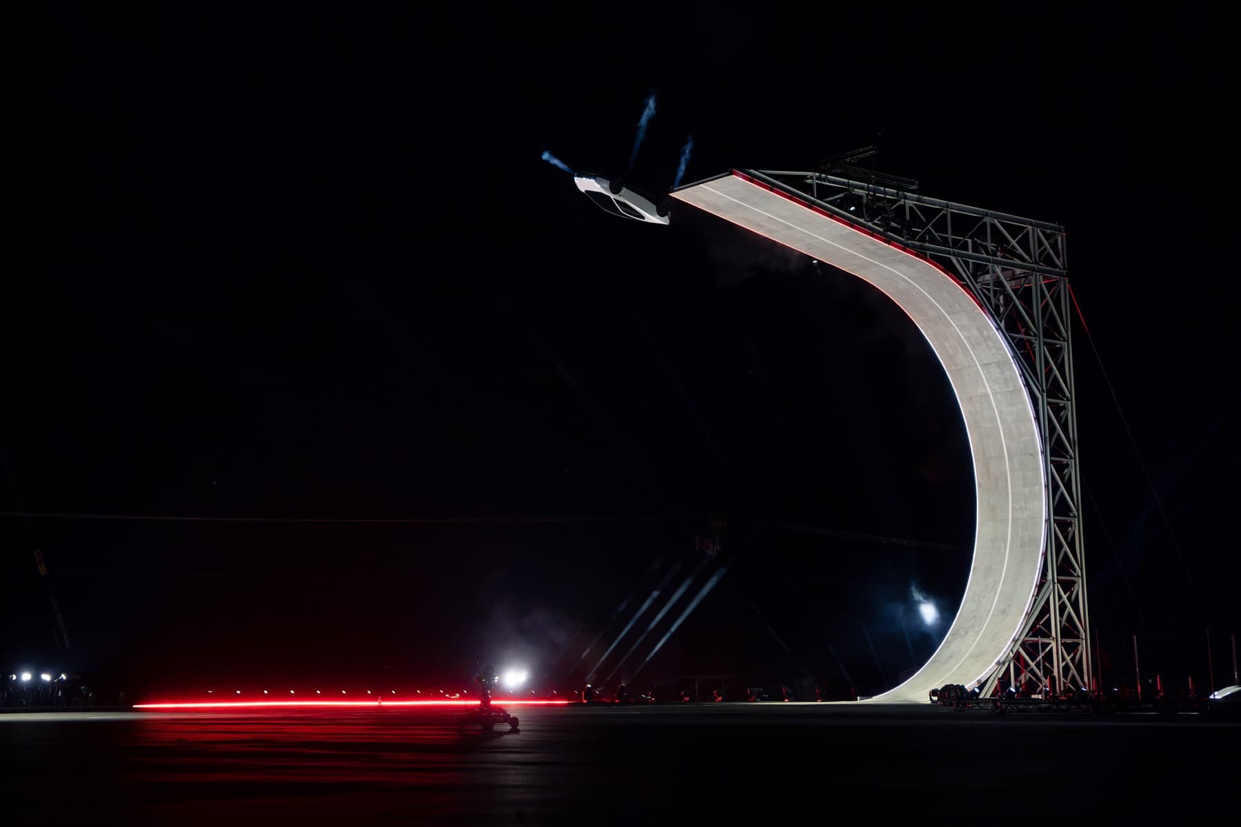 Roadster inverted just beyond a steep curved ramp, with white gas plumes visible.
